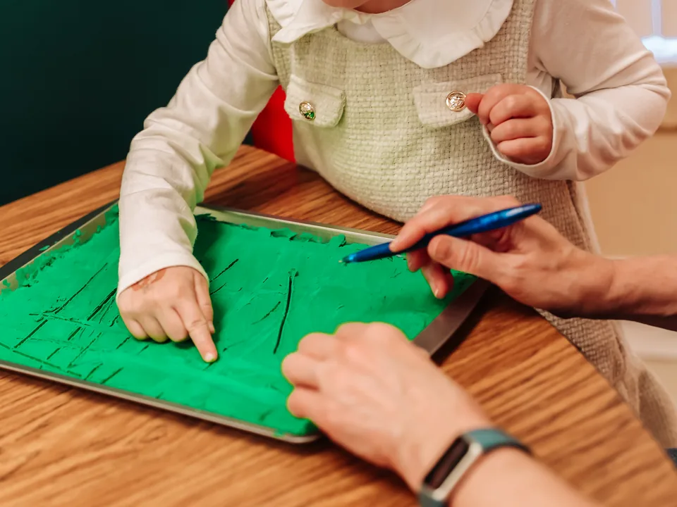 A child traces letters in a tray of green sand while a therapist demonstrates the stroke