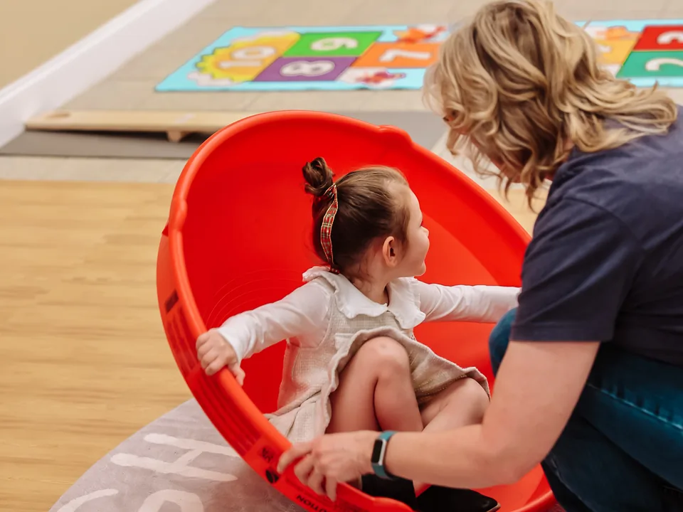 A therapist steadies a smiling child spinning in a red therapy dish on the clinic floor